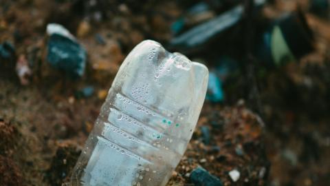 A plastic water bottle pokes out of brown soil dotted with rocks, with other plastic trash out of focus in the background.